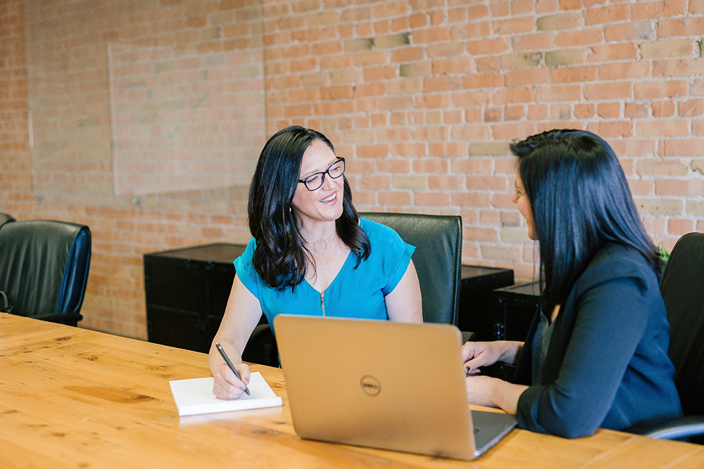 Two women sit at a wooden table in a brick-walled conference room. One woman with dark hair and glasses, wearing a blue top, smiles while writing on a notepad. The other woman, also with dark hair, in a black blazer, sits across from her, engaged in conversation. A laptop is open in front of them.
