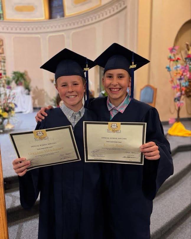 Two 6th grade boys wearing caps and gowns holding their diplomas and smiling at the camera