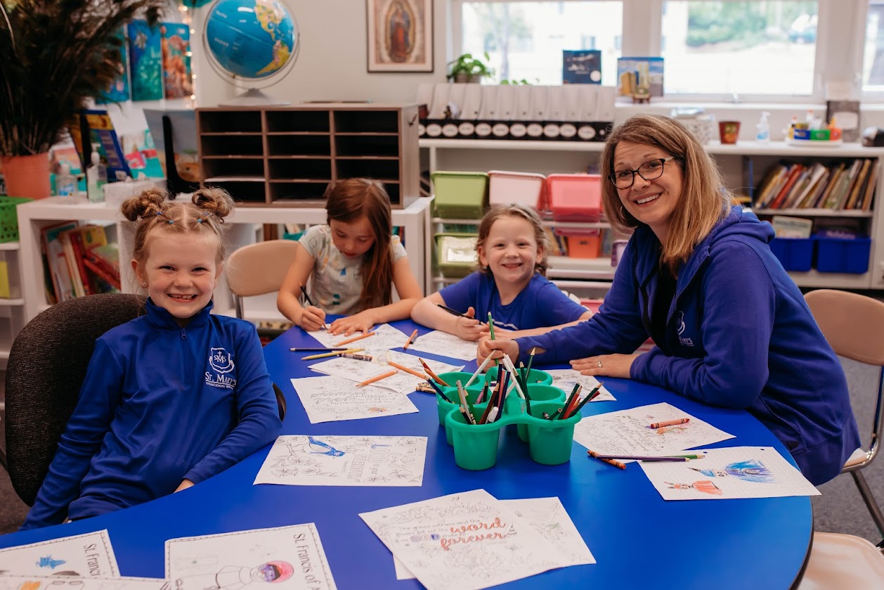 A teacher and 3 young girls sitting at a table working on a worksheet, all are looking at the camera and smiling